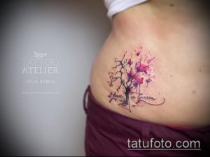 Фото тату на талии с изображением цветущей ветки сакуры, выполненное в акварельной технике с использованием розовых и фиолетовых оттенков.