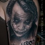 Фото тату реалистичного портрета Джокера в черно-белой гамме с надписью 'WHY SO SERIOUS?'. Рисунок выполнен на руке.