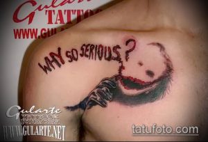 Фото тату: татуировка на плече с изображением безумной улыбки Джокера и его руки, выполненная в черно-красных тонах с эффектом брызг крови.