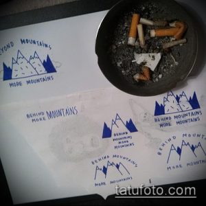 Несколько эскизов татуировок с изображением синих гор и надписью 'Beyond Mountains More Mountains'. Рядом пепельница с окурками.