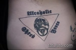 Фото простой татуировки с изображением черепа в треугольнике и надписью 'Alcoholic Shred Force', выполненной черными чернилами.