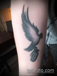 Фото тату на предплечье: силуэт ворона, держащего в лапах гранату, выполненный в черно-серых тонах.