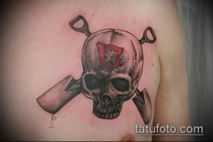 Фото тату черепа с числом 13 на лбу и двумя скрещенными лопатами, выполненное в реалистичном стиле.