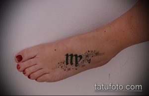 Фото тату: Небольшой символ Девы на женской стопе, украшенный тонкими линиями и брызгами краски. Изящный рисунок татуировки.