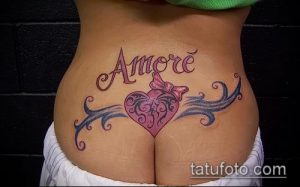 Фото тату с надписью 'Amore' и изображением сердца с бантиком на женской пояснице. Рисунок татуировки в стиле new school с яркими цветами и плавными линиями.