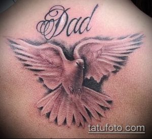 Фото тату: реалистичный рисунок летящего голубя с надписью "Dad", выполненный на спине. Черно-серые тона, детализированные перья и трогательное послание.