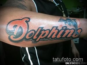 Фото татуировки на предплечье с надписью "Dolphins" и изображением дельфина. Использованы яркие цвета: красный, оранжевый, голубой и черный.