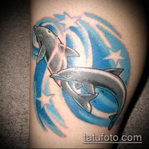 Фото тату на руке с изображением двух дельфинов, прыгающих на фоне голубых волн и ярких звезд. Рисунок выполнен в ярких, насыщенных цветах.