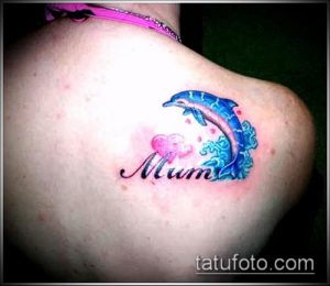 Фото тату дельфина, выпрыгивающего из воды, с надписью "Mum" и изображением сердца на плече. Изображение выполнено в ярких цветах.