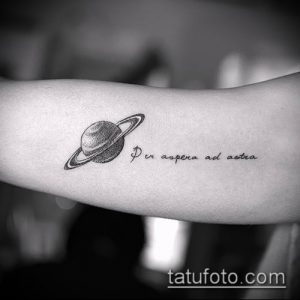 Фото небольшой татуировки Сатурна на предплечье с латинской фразой 'Per aspera ad astra'. Черно-белый стиль, реалистичное изображение планеты.
