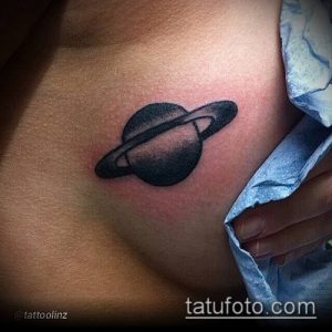 Фото небольшой татуировки Сатурна на женской груди. Рисунок выполнен в черных чернилах с использованием штриховки для создания объема. Элегантный и стильный дизайн.