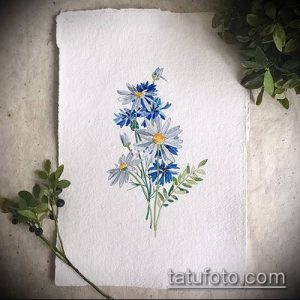 Фотография акварельной татуировки с изображением букета полевых цветов, преимущественно васильков, на белом фоне. Мягкие переходы цветов и деликатная прорисовка.
