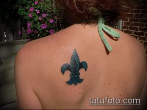 Фото небольшой татуировки флёр-де-лис, выполненной в сине-зеленых тонах, расположенной на спине девушки.