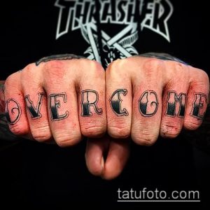 Фото тату на костяшках пальцев: надпись 'Overcome' выполнена черными чернилами в готическом стиле, создавая эффектный и мотивирующий образ.