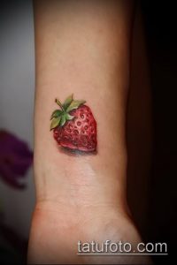 Фото маленькой татуировки клубники с зелеными листьями на запястье.