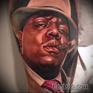 Фотография реалистичной татуировки с изображением Notorious B.I.G. в шляпе и с сигарой во рту. Детализированный портрет рэпера.