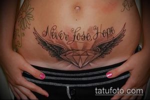 Фото тату: татуировка на животе с надписью "Never Lose Hope" и изображением крыльев. Рисунок татуировки выполнен в черно-серых тонах.