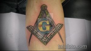 Фото тату на предплечье: масонский символ - квадрат и циркуль с буквой 'G' внутри, выполненный в серо-черных тонах.