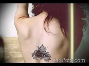 Фото тату на спине: Всевидящее Око в треугольнике с надписью 'Real Eyes Realize Real Lies'. Черно-белый рисунок.