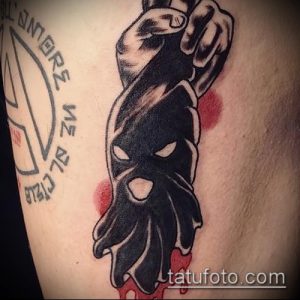Фото тату на руке: черная маска с прорезями для глаз, выполненная в стиле blackwork с использованием плотных черных заливок и минималистичных деталей.