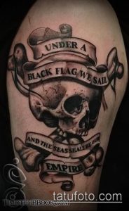 Фото тату с надписью "Under a black flag we sail" и черепом на руке. Черно-серый рисунок в стиле олдскул с четкими контурами.