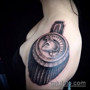 Фото татуировки на плече, изображающее шлем воина с мечом. Детализированный рисунок в черно-белых тонах с акцентом на текстуры и тени.