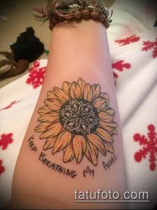Фото татуировки подсолнуха на запястье с надписью "Keep Breathing My Angel". Использованы теплые желтые и коричневые оттенки.