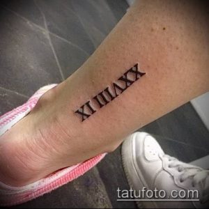 Фото тату: небольшие римские цифры 'XI IIIIAX' на женской лодыжке, выполненные черными чернилами. Изящный и лаконичный рисунок татуировки.