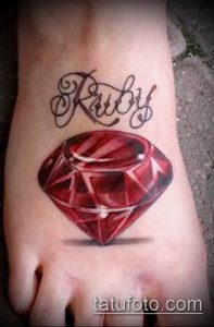 Фото татуировки рубина и надписи 'Ruby' на женской стопе. Красный драгоценный камень и черная каллиграфия создают стильный дизайн.