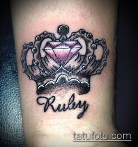 Фото тату короны с большим рубином в центре и надписью 'Ruby' под ней. Черно-серая татуировка на предплечье, детализированный рисунок.