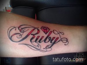 Фото тату надписи 'Ruby' с маленьким рубином над буквой 'R'. Классический рисунок татуировки с каллиграфическим шрифтом и красным камнем.