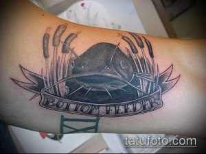 Фото тату сома на руке в стиле Old School с надписью "Bottom Feeder". Классический рисунок татуировки.