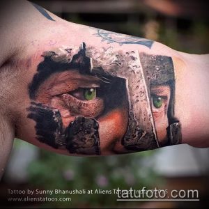 Фото тату на руке с изображением лица спартанского воина в шлеме. Реалистичный рисунок татуировки с зелеными глазами и детальной проработкой брони.