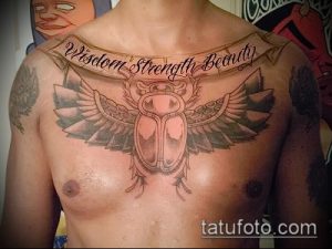 Фото тату: татуировка скарабея с расправленными крыльями и надписью "Wisdom Strength Beauty" на груди мужчины.