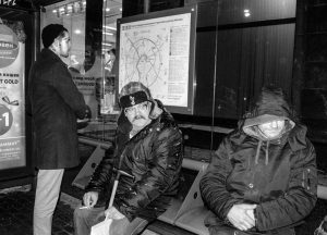 Черно-белое фото бездомного мужчины с татуировками на лице, сидящего на скамейке возле магазина. Мужчина курит сигарету.