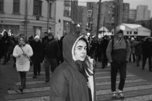Черно-белое фото девушки с татуировками на лице, переходящей дорогу на пешеходном переходе. Девушка одета в куртку с капюшоном, ее взгляд направлен в сторону.