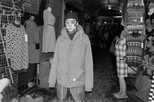 Черно-белое фото мужчины с татуировками на лице, стоящего в торговом ряду. Мужчина одет в куртку и шапку, его взгляд направлен в камеру.
