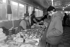 Черно-белое фото мужчины с татуировками на лице, стоящего у мясного прилавка. Мужчина одет в куртку и шапку, его взгляд направлен вниз.