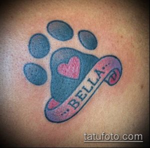 Фотография татуировки отпечатка лапы с лентой, на которой написано имя "Bella". Нежный рисунок татуировки в розовых и серых тонах.