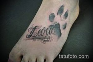 Фото татуировки на стопе с изображением отпечатка лапы и надписью "Laura". Черно-серый рисунок, каллиграфический стиль, женственная татуировка.