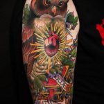 Фото татуировки на руке: сова, держащая в когтях сердце, рядом с книгами и лентой с надписью. Яркие цвета и детализированная проработка.