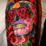 Фото татуировки в стиле Day of the Dead с изображением черепа, украшенного розами и другими цветами. Яркие цвета и детализированная проработка.