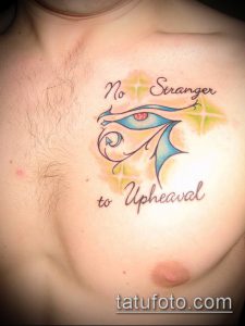 Фото татуировки Глаза Гора с надписью "No Stranger to Upheaval" на груди. Цветная татуировка с использованием синих, желтых и черных оттенков.