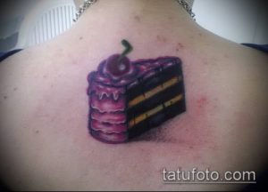 Фотография татуировки с изображением аппетитного куска шоколадного торта с вишней на спине. Реалистичный рисунок с розовой глазурью.