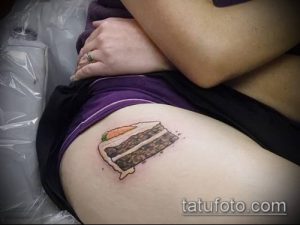 Фото тату на бедре: реалистичный кусочек шоколадного торта с кремом и апельсиновой долькой, выполненный в теплых тонах.