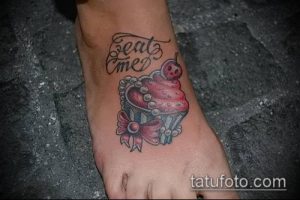 Фото тату капкейка с вишней и надписью 'Eat Me' на стопе. Оригинальный и игривый рисунок татуировки, выполненный в ярких цветах.