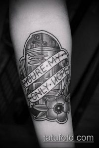 Фото тату R2-D2 на предплечье. Черно-белая татуировка в стиле олдскул с добавлением цветочных элементов и надписи.