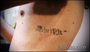 Фото татуировки с изображением группы крови AB(IV)Rh-, выполненной черными чернилами на коже.