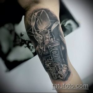 Фото тату: изображение бутылки виски Jack Daniel's, денег и шляпы, выполненное в черно-серых тонах на предплечье.