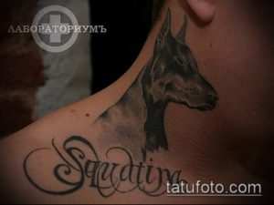 Фото черно-серой татуировки добермана на шее с подписью имени "Saadina". Реалистичный рисунок татуировки с детальной проработкой шерсти.